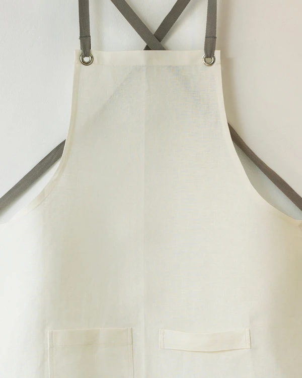 White linen apron with gray cross-back straps hanging on a plain wall